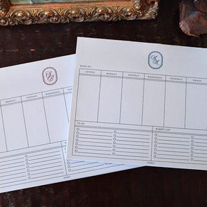 May include: Two white weekly planner sheets with black grid lines, featuring columns for Sunday through Saturday, a to-do list section, and a market list section. Each sheet displays a unique monogram at the top in a decorative border.