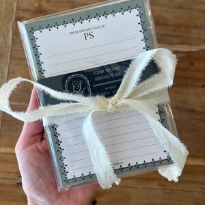 May include: A stack of lined notepads in a clear plastic sleeve, tied with a cream-colored frayed ribbon. The top sheet features a sage green border with a floral pattern, the text FROM THE KITCHEN OF: PS, and a black belly band.