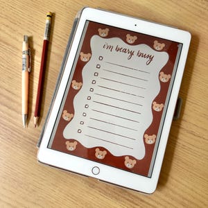 May include: A white tablet displaying a digital to-do list with a brown border featuring repeating bear faces and the text "i'm beary busy" at the top, accompanied by a light orange mechanical pencil and a red wooden pencil with a silver ferrule.