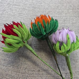 Puede incluir: Tres flores artesanales hechas de limpiapipas difusos. Una flor tiene pétalos rojos con sépalos verdes, otra tiene pétalos naranjas con sépalos verdes y la tercera tiene pétalos de color violeta claro con sépalos verdes. Cada una tiene un tallo verde.