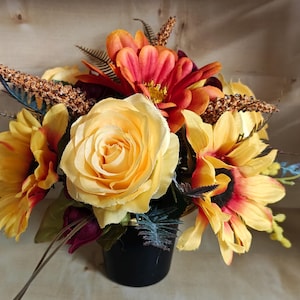 May include: A floral arrangement in a black container featuring a central yellow rose, orange and yellow sunflowers, and deep red flowers. The bouquet includes textured brown wheat-like stems and dark green fern-like foliage accents.