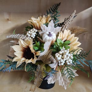Puede incluir: Un arreglo floral que presenta girasoles de color amarillo pálido, una pequeña calabaza blanca, racimos de bayas blancas y varias hojas de helecho en tonos verdes y dorados. El ramo se encuentra en una base cilíndrica oscura con texturas.