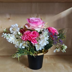 Könnte beinhalten: Ein Blumenarrangement mit einer großen zentralen rosa Rose, leuchtend rosa Nelken und kleineren weißen Blüten mit grünem Blattwerk. Die Blumen befinden sich in einer schwarzen Vase mit goldenem Rand, was einen Farbkontrast erzeugt.