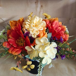 May include: A floral arrangement featuring artificial flowers in shades of orange, red, yellow, and cream. The bouquet includes large orange and red blossoms, pale yellow petals, small purple accents, and green fern-like foliage in a black container.