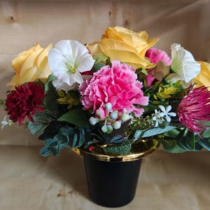 Puede incluir: Un arreglo floral con rosas amarillas, flores de campanilla blancas, un clavel rosa brillante central y crisantemos rojos oscuros. El ramo incluye follaje verde y pequeñas bayas de color verde claro, todo en un florero negro con borde dorado.