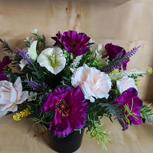 May include: A floral arrangement in a black pot featuring deep purple poppies, pale pink roses, and white lilies. The bouquet is accented with sprigs of purple lavender, green foliage, and small yellow filler flowers, creating a dense, colourful display.