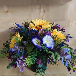 May include: A floral arrangement in a black container featuring bright yellow sunflowers, purple morning glory flowers with white centres, and various green foliage. The bouquet includes small purple filler flowers and textured greenery.