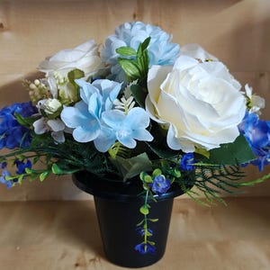 May include: A floral arrangement in a black plastic pot featuring white roses, light blue hydrangea-like blossoms, and smaller dark blue flowers. The bouquet includes green foliage and stems, creating a rounded display of mixed blue and white colours.