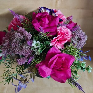 May include: A floral arrangement featuring vibrant magenta roses, soft pink carnations, and clusters of purple filler flowers. The bouquet includes green foliage and small white blossoms, creating a dense, rounded display of artificial botanical elements.