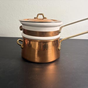 Pode incluir: Um recipiente para banho-maria composto por uma panela de base em cobre polido com duas pequenas pegas laterais e um cabo longo em latão. O encaixe superior é em esmalte branco com uma faixa de cobre e uma tampa de cobre a condizer com pega em latão.
