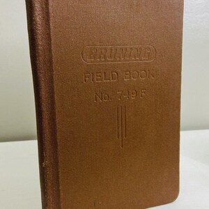 May include: A brown, rectangular field book with a textured cover. The front cover features the embossed text BRUNING FIELD BOOK No. 749 F, with three vertical lines below the text. The book has a rounded spine and a sturdy, bound construction.