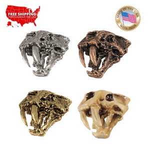 May include: Four different colored metal and bone-colored replicas of a saber-tooth cat skull. The skulls are facing forward with their mouths open and their fangs showing. The colors are silver, bronze, gold, and bone.
