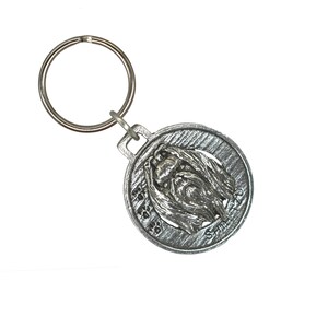 May include: Silver keychain featuring a detailed image of a Shih Tzu dog's face. The keychain has a round metal charm with a textured surface and a silver key ring.