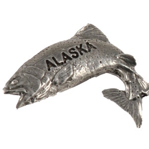 May include: A silver-colored metal fish figurine with the word "ALASKA" engraved on its side. The fish is jumping out of the water with its mouth open.