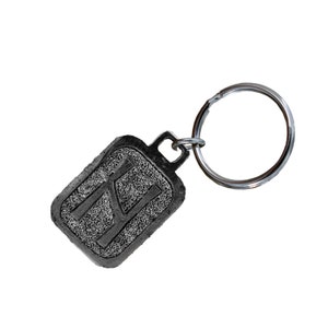 May include: A silver keychain with a rectangular tag featuring the letter "K" in a stylized font. The tag has a textured surface.