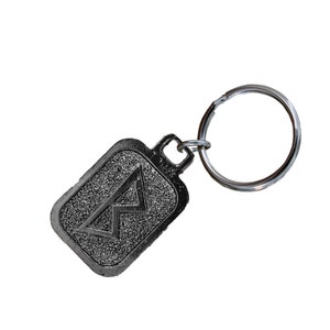 May include: A silver-colored keychain with a rectangular tag featuring a raised, textured, black rune symbol. The keychain has a silver-colored ring.