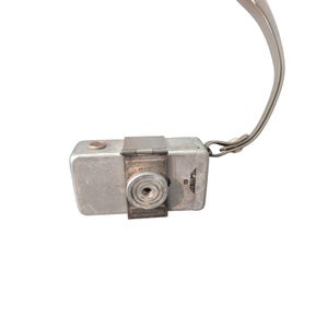May include: A rectangular silver-toned metal subminiature camera featuring a central circular lens housing, a top-mounted shutter button, and a thin grey wrist strap attached to the side. The camera body has a matte finish with a hinged metal plate.