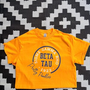 May include: A bright orange short-sleeve t-shirt featuring a circular blue and white graphic. The text reads SIGMA GAMMA RHO, BETA TAU, 1922, and Pretty Poodles in a script font. The design is centered on the chest of the garment.