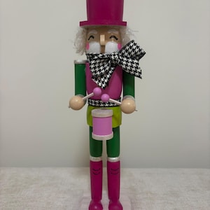 May include: A wooden nutcracker figure wearing a bright pink top hat and boots, a green jacket, and a pink waistcoat. It features a black and white houndstooth bow tie, white hair, and holds two small pink drumsticks over a pink drum on its chest.