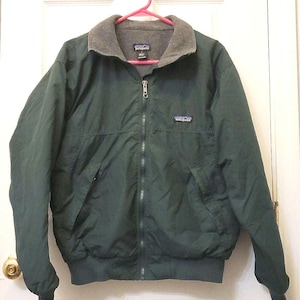 Puede incluir: Una chaqueta Patagonia de color verde oscuro con cremallera frontal completa, cuello forrado de forro polar gris y mangas largas con puños acanalados. La chaqueta presenta un pequeño logotipo de la marca Patagonia en el pecho izquierdo.