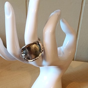 Könnte beinhalten: Ein silberner Löffelring mit ovaler Oberfläche und aufwendigen Details. Der Ring wird auf einer weißen Keramikhand präsentiert, die das Design und die Handwerkskunst hervorhebt. Der Ring ist ein einzigartiges Schmuckstück.