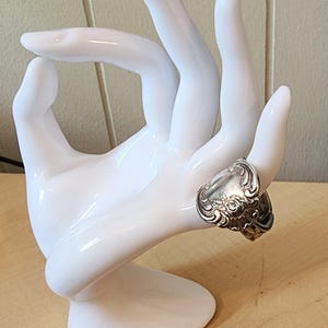 May include: A silver-toned ring with ornate floral detailing is displayed on a white hand-shaped jewelry holder. The ring's design features intricate patterns and a vintage aesthetic, showcasing a unique piece of jewelry.