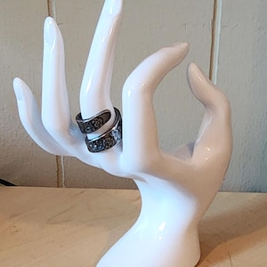 May include: A white ceramic hand-shaped ring holder displaying two silver rings with floral designs. The rings are wrapped around the fingers of the hand. The holder is on a light-colored wooden surface.