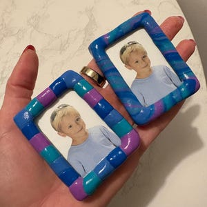 May include: Two rectangular picture frames crafted from marbled polymer clay in shades of blue, purple, and teal. Each frame holds a small photograph of a young child with blonde hair wearing a light blue shirt and sunglasses on their head.