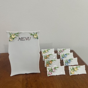 May include: A white ceramic menu holder and seven matching place card holders, each featuring delicate hand-painted floral accents in yellow, pink, and blue with green leaves. The menu holder displays the word MENU in black lettering.