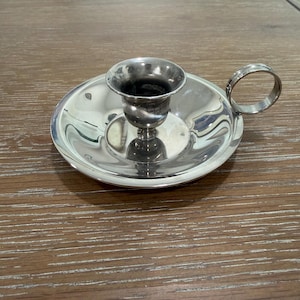May include: A polished silver-toned metal candle holder featuring a wide, circular drip tray base and a central cup for holding a candle. A single, circular finger loop handle is attached to the side of the tray, allowing for easy transport.