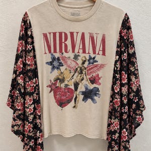 May include: A cream-colored t-shirt featuring the red text NIRVANA and a graphic of a winged figure surrounded by blue and yellow flowers and a red heart. The shirt has long, loose black sleeves with a repeating red and white floral pattern.