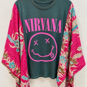May include: A dark green t-shirt featuring the word NIRVANA and a pink smiley face graphic. The shirt has wide, flowing sleeves made from a quilted pink fabric with a floral pattern in shades of blue, brown, and cream, hanging from a hanger.