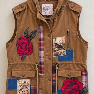 May include: A brown hooded vest featuring a Rebel Bloom label, decorated with red floral patches, plaid fabric strips, and rectangular patches depicting birds. The garment includes a front zipper, snap buttons, and two front pockets with flaps.