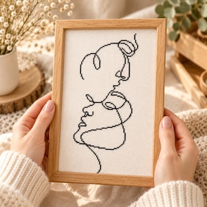 May include: A framed cross-stitch artwork featuring a continuous black line drawing of two faces in profile, one above the other, stitched onto white fabric. The piece is enclosed in a light-toned wooden frame with a simple, clean rectangular border.