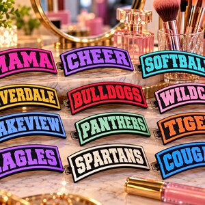 May include: A collection of twelve rectangular hair clips with curved edges, each featuring bold varsity-style lettering in various colors including pink, purple, blue, yellow, red, orange, green, and black, displaying words like MAMA, CHEER, SOFTBALL, RIVERDALE, BULLDOGS, WILDCATS, LAKEVIEW, PANTHERS, TIGERS, EAGLES, SPARTANS, and COUGARS.