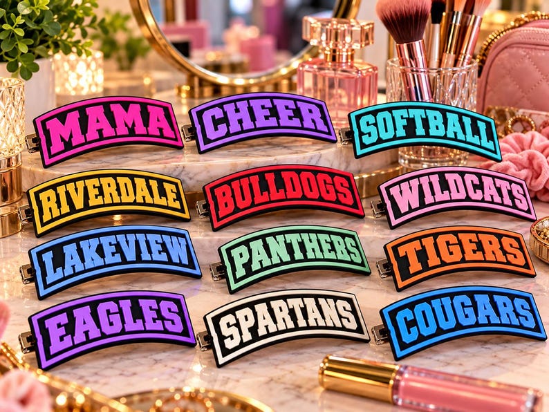 May include: A collection of twelve rectangular hair clips with curved edges, each featuring bold varsity-style lettering in various colors including pink, purple, blue, yellow, red, orange, green, and black, displaying words like MAMA, CHEER, SOFTBALL, RIVERDALE, BULLDOGS, WILDCATS, LAKEVIEW, PANTHERS, TIGERS, EAGLES, SPARTANS, and COUGARS.