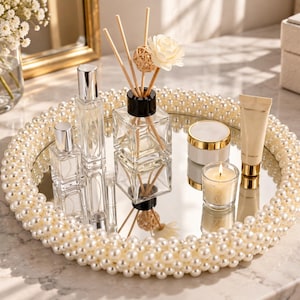 May include: A round mirrored vanity tray featuring a thick border of layered white faux pearls. The tray holds two clear glass perfume bottles with silver caps, a square reed diffuser with wooden sticks, a small white jar, and a tube of cream.