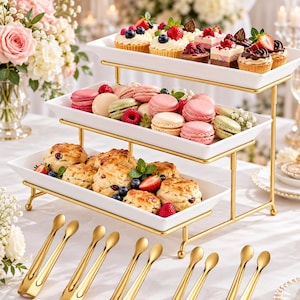 May include: A three-tiered white rectangular serving tray set with a gold metal frame. The trays hold assorted cupcakes, macarons, and scones topped with berries. Five pairs of gold-toned metal serving tongs are arranged in the foreground.