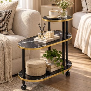 May include: A three-tier rolling side table featuring oval shelves with a black marble-patterned surface and gold-toned metal edges. The unit is supported by black cylindrical legs and includes small wheels at the base for mobility.
