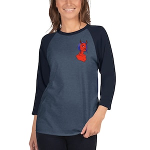 Puede incluir: Una camiseta de béisbol azul marino y gris jaspeado con un gráfico de diablo rojo en el pecho. El gráfico es una ilustración estilizada de un diablo femenino con piel roja, cuernos y cabello azul. Las mangas son azul marino y el cuerpo es gris jaspeado.