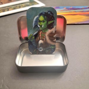 May include: A miniature painting of a green-skinned orc warrior with an axe, displayed inside a small, open, silver-colored tin. The orc is wearing brown armor and a fur-like shoulder piece. The background is a mix of blues and greens.