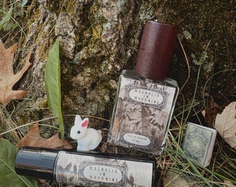 Può includere: Un flacone di profumo rettangolare in vetro con tappo marrone scuro e un flacone cilindrico roll-on, entrambi etichettati Magnolia in Mourning, accanto a una piccola statuetta di coniglio bianco e un libro in miniatura.