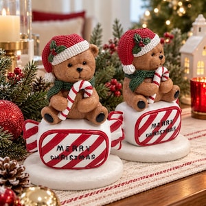 Pode incluir: Duas figuras de ursos de peluche castanhos com gorros de malha vermelhos com rebordo branco e cachecóis verdes. Cada urso segura um bastão de rebuçado vermelho e branco sobre uma base branca em forma de rebuçado às riscas com Merry Christmas.