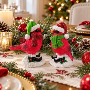 Pode incluir: Duas figuras de pássaros cardeais vermelhos sobre bases cobertas de neve. O pássaro da esquerda usa um gorro verde e cachecol vermelho, enquanto o da direita usa gorro vermelho e cachecol verde. Ambos têm máscaras pretas e bicos laranja.