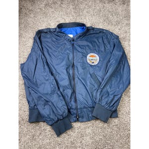 Puede incluir: Una chaqueta cortavientos azul marino con cremallera, cuello alzado y puños acanalados. La prenda presenta un parche circular en el pecho izquierdo que muestra un paisaje desértico con el texto "U.S. BUREAU OF RECLAMATION" y "HOOVER DAM STAGE II".