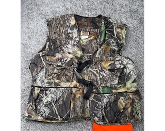 Op de afbeelding: Een mouwloos vest met een bruin, groen en zwart camouflagepatroon, voorzien van meerdere voorzakken, zwarte gespsluitingen en een Born Afield-merklabel. Een apart feloranje stuk stof is onder het vest geplaatst.