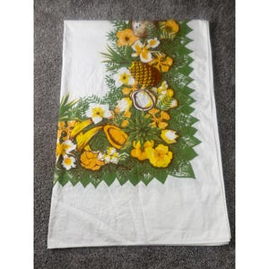 May include: A white rectangular tablecloth featuring a vibrant corner print of tropical fruits and flowers. The design includes yellow hibiscus blossoms, green palm fronds, pineapples, and sliced coconuts arranged in a dense, leafy border.