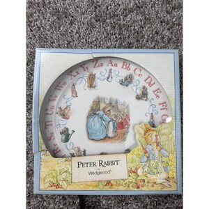 May include: A white ceramic plate featuring Peter Rabbit illustrations, the alphabet in red letters, and blue scrollwork. The plate is housed in a square cardboard box with a Peter Rabbit graphic and the text Peter Rabbit by Wedgwood.