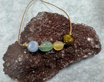 May include: A bracelet featuring four smooth, polished gemstone beads in light blue, pale green, translucent yellow, and brown tiger's eye, strung on a thin, adjustable tan cord with small gold-toned metal accents at the ends.