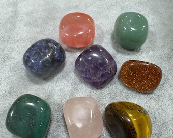 May include: A collection of eight polished, smooth, tumbled stones in various colours including a translucent pink, a deep purple, a dark blue with black flecks, a shimmering gold-brown, a light green, a dark green, and a soft pale pink.